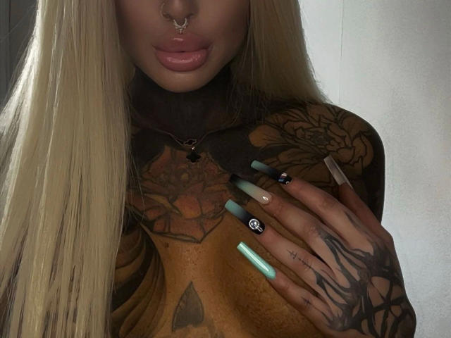 Lexxis-Blck, uma mulher atraente com cabelos loiros longos, piercing no nariz, lábios rosados brilhantes, tatuagens no peito e mãos com unhas verdes, azuis e pretas, exibindo suas tatuagens e seios..