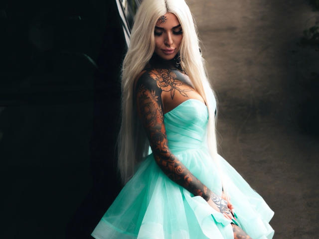 Lexxis-Blck, met lang, platina blond haar, tatoeages op haar armen en borst, draagt een groene, strapless jurk van tule, staat met een zachte glimlach en kijkt naar beneden..