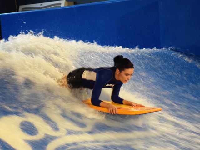 OrianaLaFrancaise è accovacciata su una tavola da surf arancione, mentre cavalca un'onda blu, indossando una muta blu e bianca, con l'acqua che le schizza intorno, e appare concentrata..