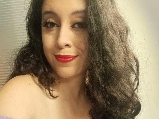 KendraSimons heeft golvend zwart haar, rode lippenstift, en ze glimlacht naar de camera met een subtiele knipoog. Haar blote schouders zijn zichtbaar, wat een sexy en zelfverzekerde uitstraling laat zien..