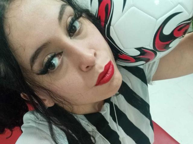 Kendra Simons, mit rotem Lippenstift, einem schwarz-weiß gestreiften Hemd und einem Fußball vor dem Gesicht, hat lockiges, schwarzes Haar und intensive, grüne Augen und blickt nach oben in die Kamera..