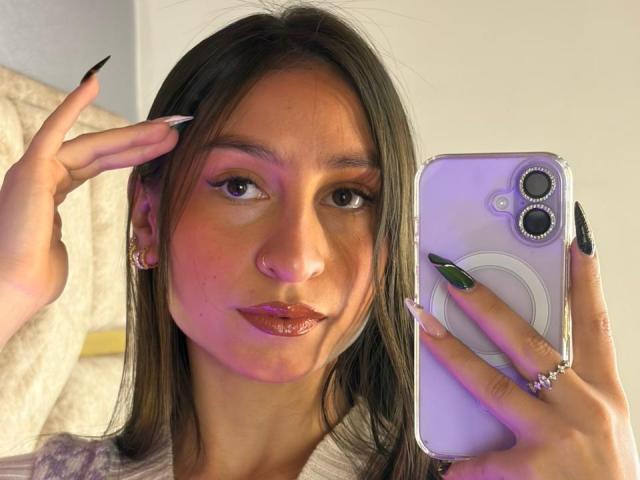 MaddyCarina with long brown hair, pink eyeshadow, glossy lipstick, black nails, holding a purple iPhone, making a peace sign, wearing a ring, nose ring, and earrings.