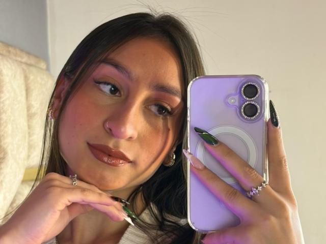 MaddyCarina with long brown hair, holding a purple phone, showing off green and black nail polish, and wearing earrings, rings, and a choker. She has pinkish eyeshadow and glossy lips.
