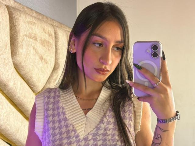 MaddyCarina, com cabelos longos e pretos, pele clara, vestindo um suéter roxo e branco, tirando uma selfie com um celular roxo, unhas verdes, um piercing no nariz e uma pulseira no pulso..