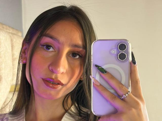 MaddyCarina with dark brown hair, green eyes, and glossy red lips, holding a pink phone, wearing a ring and black nails, has a nose ring and subtle makeup.