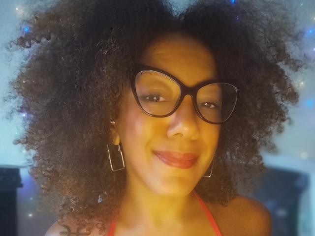 CinnamonBabe ha i capelli neri e ricci, indossa occhiali neri e ha un sorriso delicato con rossetto rosso. Porta orecchini quadrati e un top a halter rosso..