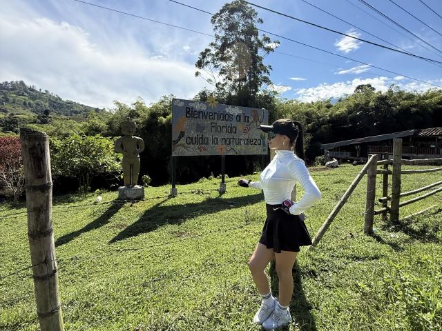 KimmyK se tient debout sur l'herbe, vêtue d'un pull blanc à col roulé, d'une jupe noire, de baskets blanches et d'une casquette noire, avec ses fesses légèrement visibles, près d'un panneau en bois et d'une statue..