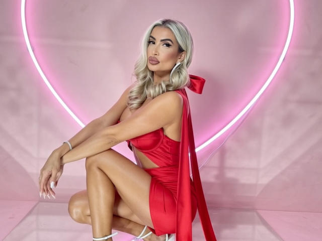 Lellie with blonde hair, red dress, and big bow, squatting, showing her ass, in white heels, pink neon heart, shiny skin, and a sexy, confident look.
