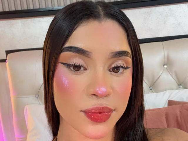 LunaVali has shiny pink cheeks, thick black eyeliner, long eyelashes, glossy red lips, and straight dark brown hair. Her tan skin and brown eyes are visible.