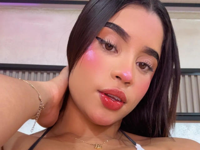 LunaVali's face with pink blush, glossy red lips, long dark hair, and a gold necklace, holding her hair back, wearing a chain bracelet, looking slightly seductive.