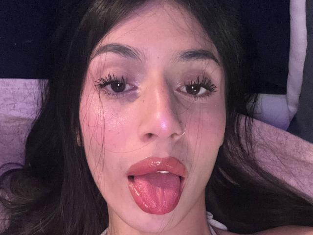 SophieBeaumont with dark brown hair, nose ring, and slightly glossy pink lips, tongue out, staring at camera with heavy eyelashes and subtle makeup. Her face is slightly shiny.
