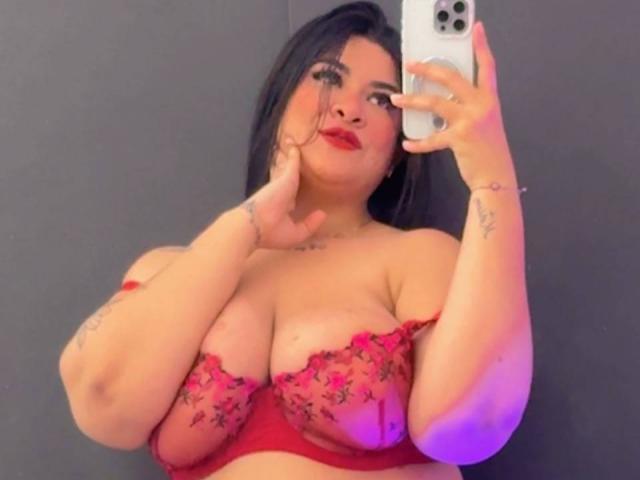 Rashael: Grande, con reggiseno rosso in pizzo, che mostra un seno prosperoso, tiene il telefono allo specchio, capelli scuri, rossetto rosso, tatuaggi sul braccio, guarda la fotocamera, sfondo grigio..