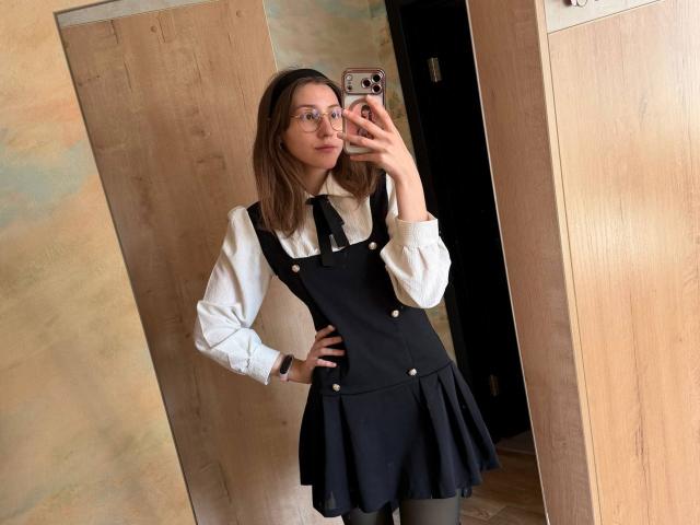 Lenore Schontz, portant des lunettes, est vêtue d'une robe noire à bretelles portée par-dessus une chemise blanche, elle tient un téléphone, se tient devant un miroir, une main sur la hanche, une porte en bois est visible en arrière-plan..