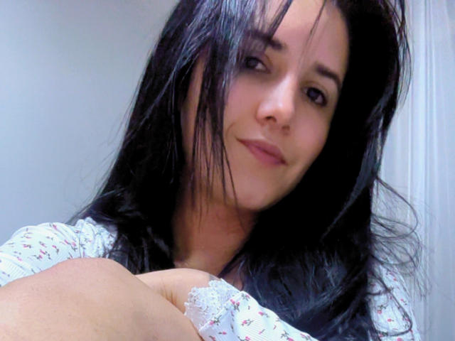 Kkatalina, con i capelli neri, la pelle chiara e un sorriso delicato, indossa una camicia a fiori, con le braccia incrociate, mostrando un piccolo dettaglio in pizzo sulla sua camicia..