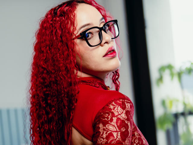 LilJane, com cabelo ruivo encaracolado, óculos pretos, blusa vermelha com renda e batom vermelho, olhando por cima do ombro, mostrando o bumbum, em um ambiente interno bem iluminado..
