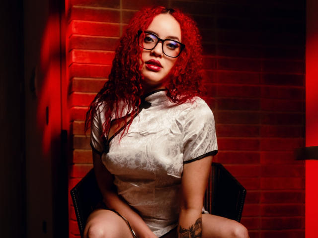 LilJane mit lockigem, rotem Haar, Brille und einem engen, weißen Kleid, sitzt auf einem Stuhl, zeigt ihren tätowierten Arm und hat leicht geöffnete Lippen, im Hintergrund ist rotes Licht..