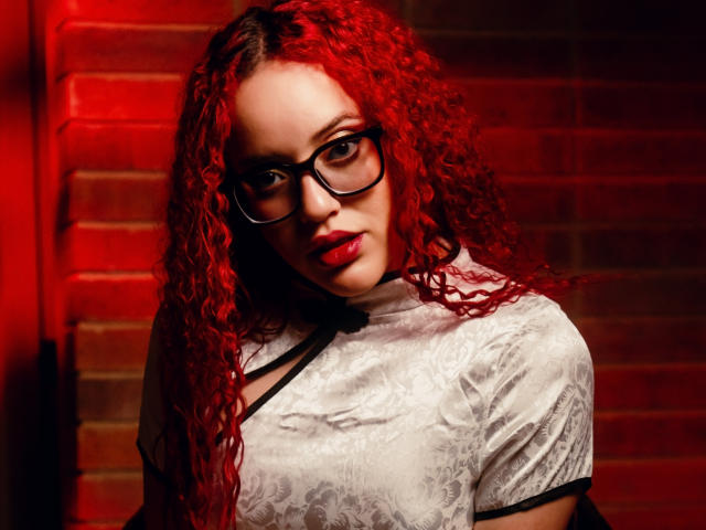 LilJane with red curly hair, wearing glasses, white lace top, black straps, and red lipstick, looking seductively at the camera, showing off her tits and a cute face.