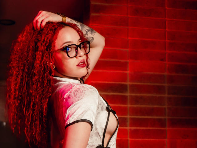 LilJane, com cabelo ruivo encaracolado, óculos e uma blusa branca com cadarços, mostrando seus seios, uma tatuagem no braço e batom vermelho, em frente a uma parede de tijolos iluminada por luz vermelha..