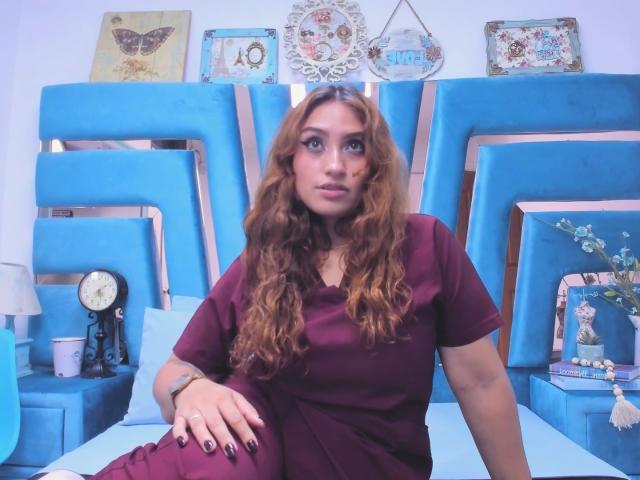 LaylaCollins with long, wavy red hair, wearing a maroon dress, sitting on a blue bed, looking up, has dark nail polish, and a ring on her right hand.