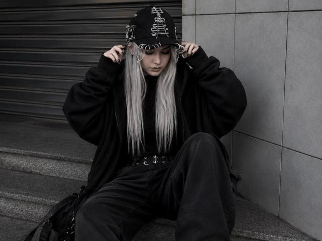 ErgoProxy, com cabelos longos e prateados, usando um boné preto com correntes, um casaco preto grande e calças pretas, sentada em degraus de pedra cinza, com as mãos ajustando o boné..