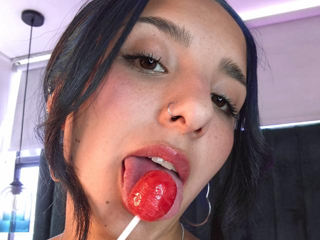 ArisSuarez with dark hair, nose ring, and hoop earrings, tongue out, licking a shiny red lollipop, eyes half-open, eyebrows raised, in a bright room.