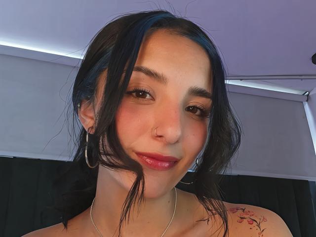 Aris Suarez, avec ses cheveux foncés, ses boucles d'oreilles, son piercing au nez et son rouge à lèvres rose, affiche un sourire discret. Elle a un tatouage sur son épaule droite et porte un collier en argent..