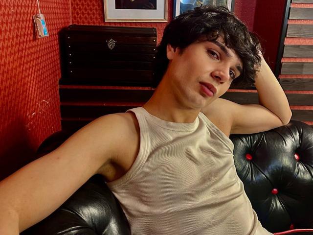 RubyMote, com cabelo preto encaracolado, vestindo uma camiseta branca, sentada em um sofá de couro preto com detalhes, com o braço apoiado na cabeça, olhando para a câmera com os lábios levemente franzidos..