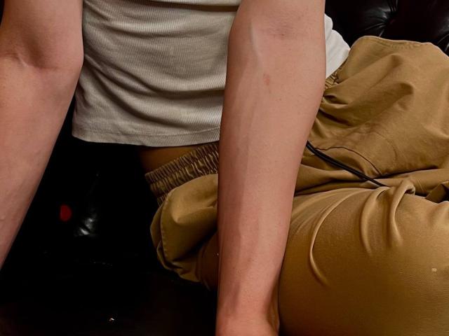 RubyMote's pale arms, wearing a white shirt, sit with brown pants, showing a slightly dirty, relaxed thigh. Black leather couch in background. 