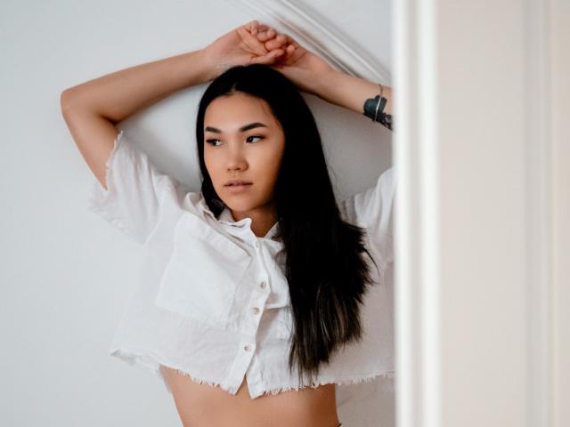 DivineMadness with long black hair, arms raised, wearing a white, frilled, button-up shirt, exposing her midriff, looking to the side with a serious expression, tattoo on right arm.