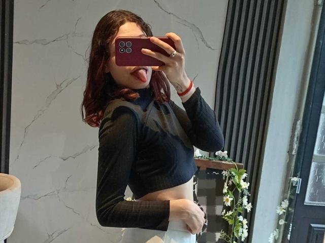 Sophie Blake, avec des cheveux roux, un haut en mesh noir, la langue tirée, tenant un téléphone devant un miroir, montrant son ventre et un pantalon blanc, portant un bracelet rouge..