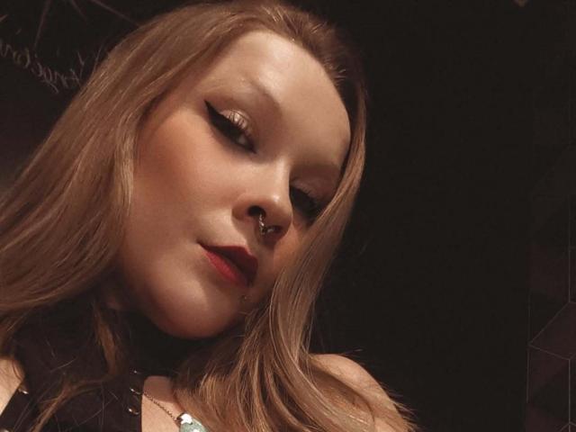 AnnaFalconeX with long blonde hair, bold eyeliner, red lipstick, nose ring, and slight smirk, wearing black choker and necklace, in dim light. Boobs not visible.