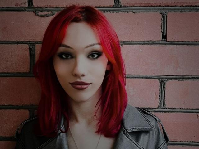 KiranaHayes with bright red hair, dark eyeliner, and red lipstick, wearing a gray jacket, has a serious expression, and a slim face, showing her tits slightly.