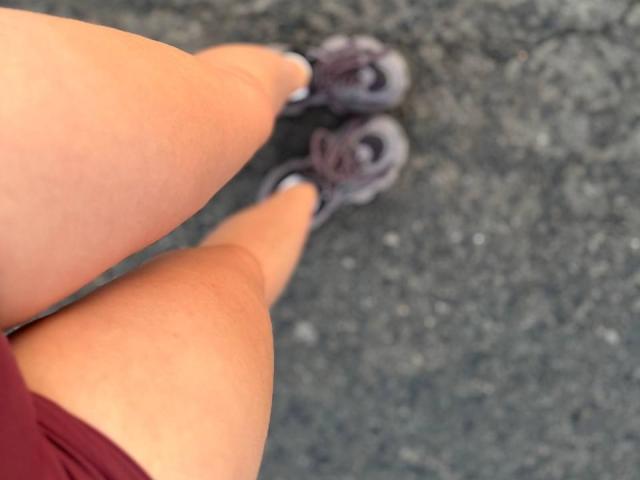 AlexXas' legs and gray sneakers are in focus, with their tan, curvy thighs and red shorts visible. The image is a top-down shot, showing a textured gray floor.