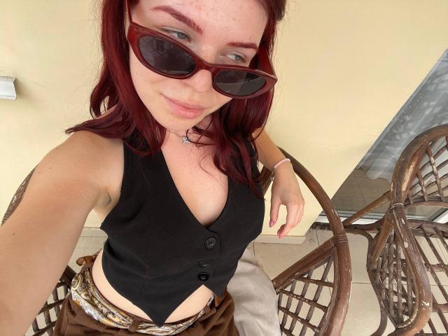 AlexXas, con i capelli rossi, indossa occhiali da sole scuri, un top nero senza maniche e pantaloni marroni, mostrando il suo seno piccolo, ed è in piedi accanto a delle sedie in vimini..