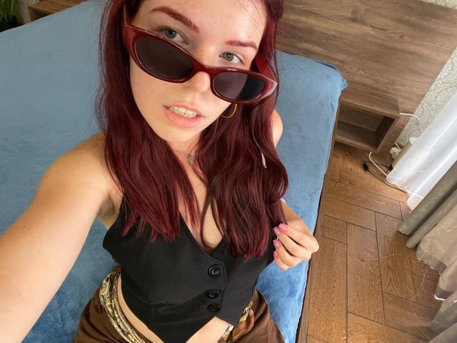 AlexXas, con cabello rojo, usando gafas de sol rojas, una camisa negra abotonada, mostrando senos pequeños, pantalones marrones y un cinturón de estampado de leopardo, en un dormitorio con piso de madera y una cama azul..