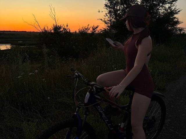 AlexXas, mit rotem Kleid, Mütze und Fahrrad, sitzt auf einem Fahrrad bei Sonnenuntergang, hält ein Telefon und zeigt ihren Hintern, in einer grünen, dunkler werdenden Landschaft..