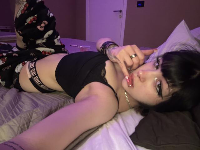 AshleyAlt è sdraiata su un letto, indossa una maglia di pizzo nero e biancheria intima "PINK", con pigiami neri, e tocca le labbra con un dito, guardando la telecamera..
