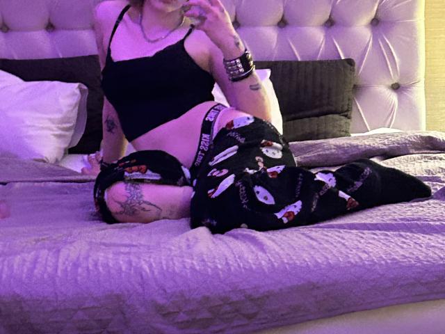 AshleyAlt, vêtue d'un débardeur noir et d'un pantalon de pyjama noir avec des motifs de personnages de dessins animés blancs, est assise sur un lit, montrant ses tatouages, sous un éclairage violet. Ses fesses sont légèrement relevées..