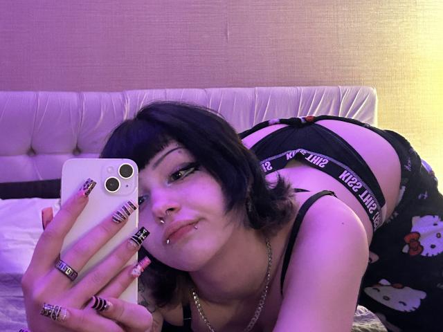 AshleyAlt, con capelli neri, piercing e trucco, è piegata in avanti, indossando biancheria intima nera, e sta scattando un selfie con un telefono bianco, mostrando il sedere e i seni, in una stanza illuminata di viola..