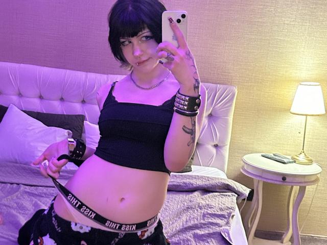 AshleyAlt, avec des cheveux noirs courts, un crop top noir et des sous-vêtements noirs avec une ceinture "Bitch Bank", tenant un téléphone, montrant son ventre nu, avec des tatouages sur le bras, dans une chambre à coucher avec une lampe..