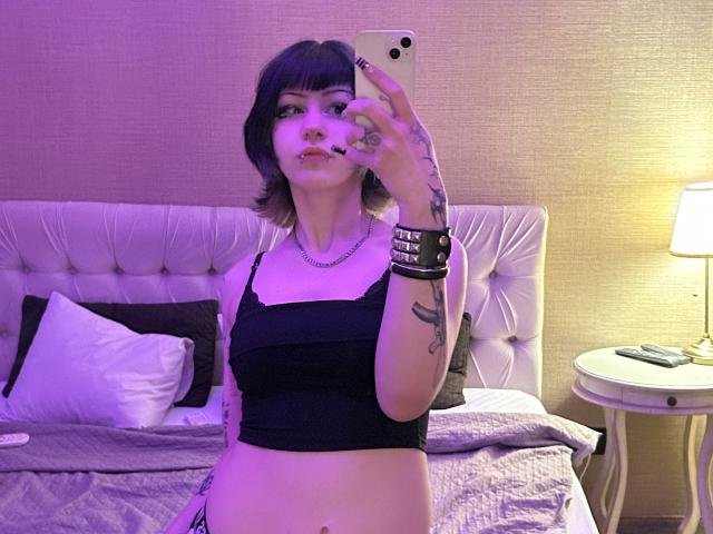 AshleyAlt, avec des cheveux violets, un crop top noir, des tatouages sur les bras, tenant un téléphone pour un selfie devant un miroir, chambre violette, tête de lit blanche capitonnée, lampe violette, table ronde blanche, lit gris..