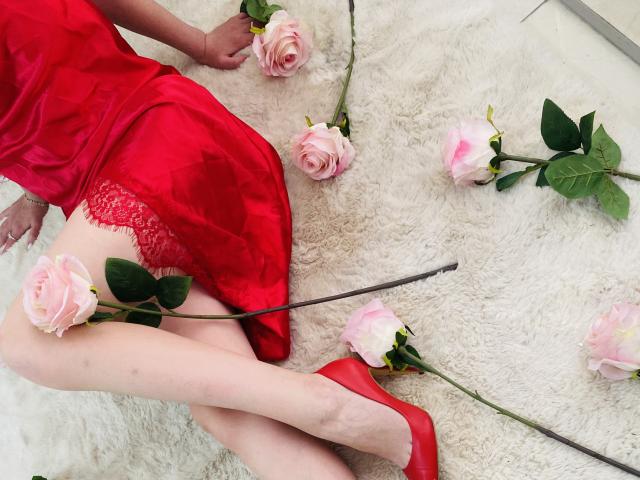 StellaNoir in red lace lingerie and satin dress, lying on white fur with pink roses, red heels, and green leaves, showing her pale thighs and red lace panties.