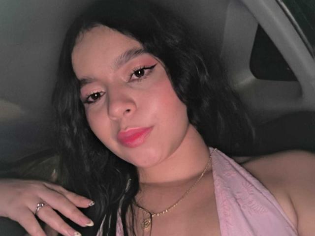 Lissy Carter hat dunkles, welliges Haar, pinken Lippenstift und eine goldene Halskette. Sie trägt ein pinkes Top mit Halstuch und einen Ring an ihrer rechten Hand und sitzt in einem Auto..