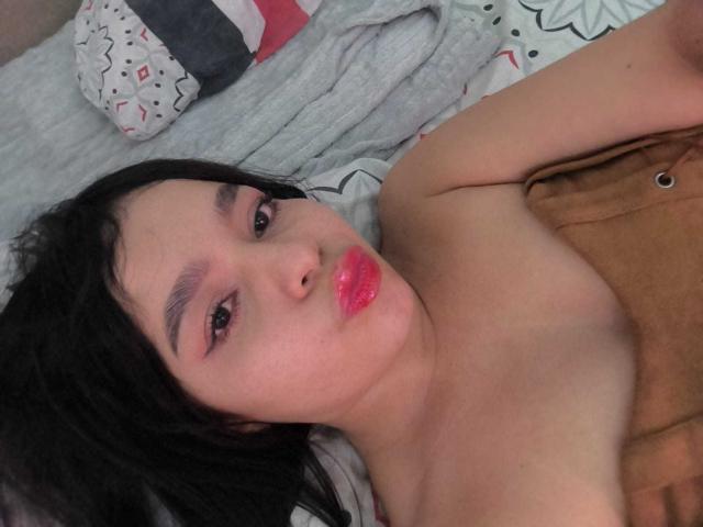 Lissy Carter mit schwarzen Haaren, pinkem Lippenstift und entblößten Schultern, liegt da, verzog das Gesicht und zeigt ihre glänzenden, rosafarbenen Lippen, trägt braune Hosen und ein graues Hemd, das teilweise sichtbar ist..
