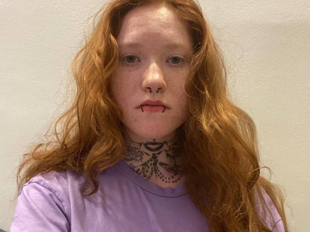 Machelle Trebesch heeft rood, golvend haar, sproeten, een neusring en een lip-piercing, en draagt een paars shirt met een gedetailleerde zwarte tatoeage op haar nek..