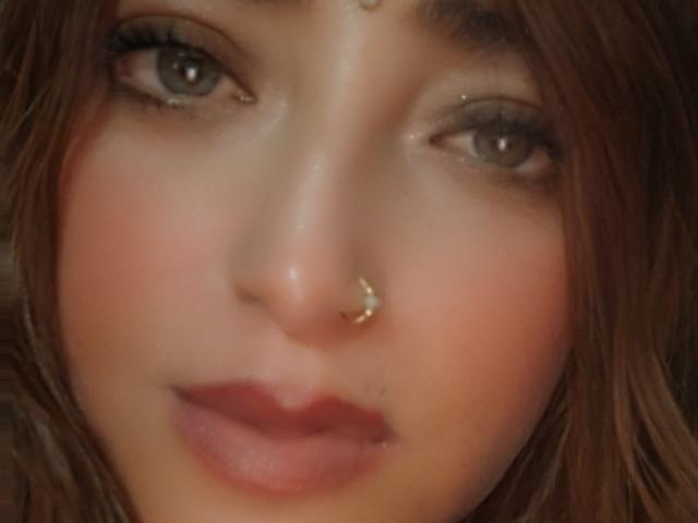 Le visage d'AlessadraJoness, avec ses yeux marron, sa peau rosée, son petit nez orné d'un anneau en or, et ses lèvres légèrement entrouvertes d'une teinte rosée. Ses cheveux bruns sont ondulés et tombent autour de son visage..