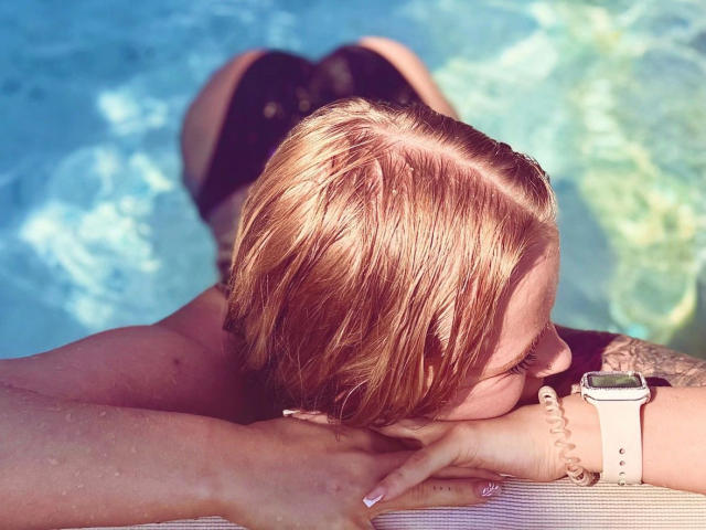 Angelxxx1- morena con cabello rubio, con los brazos apoyados en el borde de la piscina, la cara en el agua, vistiendo parte de bikini negro, un reloj rosa y una pulsera blanca, con un tatuaje en el brazo derecho..