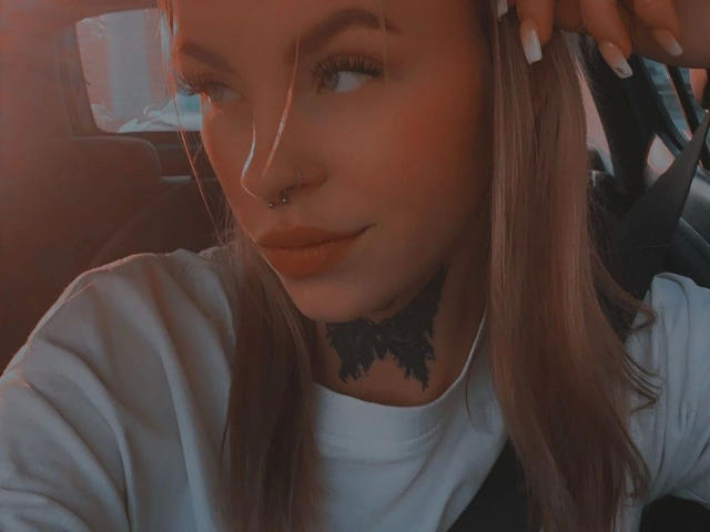 Angelxxx1, aantrekkelijk met een neusring, een tatoeage onder de nek, lang blond haar, een wit shirt, en met een hand in haar haar, zittend in een auto, naar de zijkant kijkend..