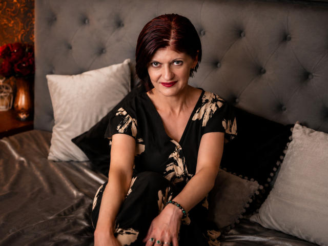 MichelleNoir, con i capelli rossi corti, seduta su un letto con un abito nero a fiori, sorridente, che mostra il seno, con lo smalto per unghie verde e un braccialetto, in una camera da letto accogliente..