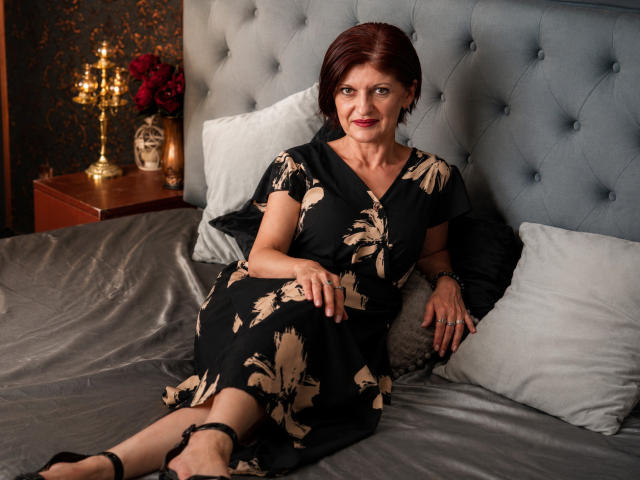MichelleNoir, eine Frau mittleren Alters mit kurzen, roten Haaren, sitzt auf einem Bett in einem schwarzen Kleid mit Blumenmuster, wobei ihre Brüste sichtbar sind, mit einem ruhigen Lächeln, schwarzen Absätzen und grauen Kissen..